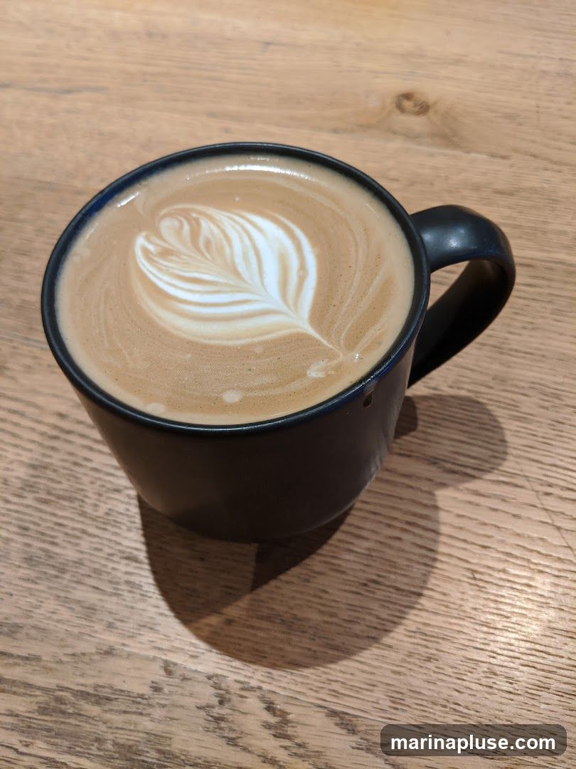 A dark mug filled with a latte, featuring intricate latte art on top. The mug is placed on a wooden surface, casting a shadow.