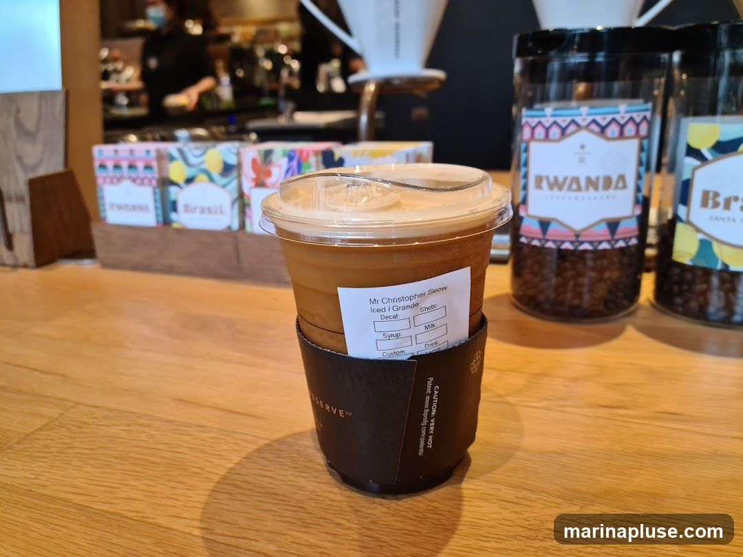 An iced coffee drink in a clear plastic cup with a lid sits on a wooden counter. The cup has a dark brown sleeve with