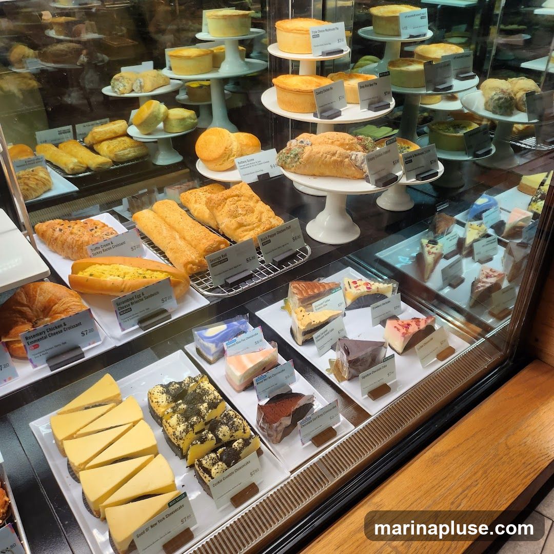A display case filled with various pastries, cakes, and savory items. Items include croissants, pies, quiches, sausage rolls, and an assortment of cheesecakes and layered cakes.