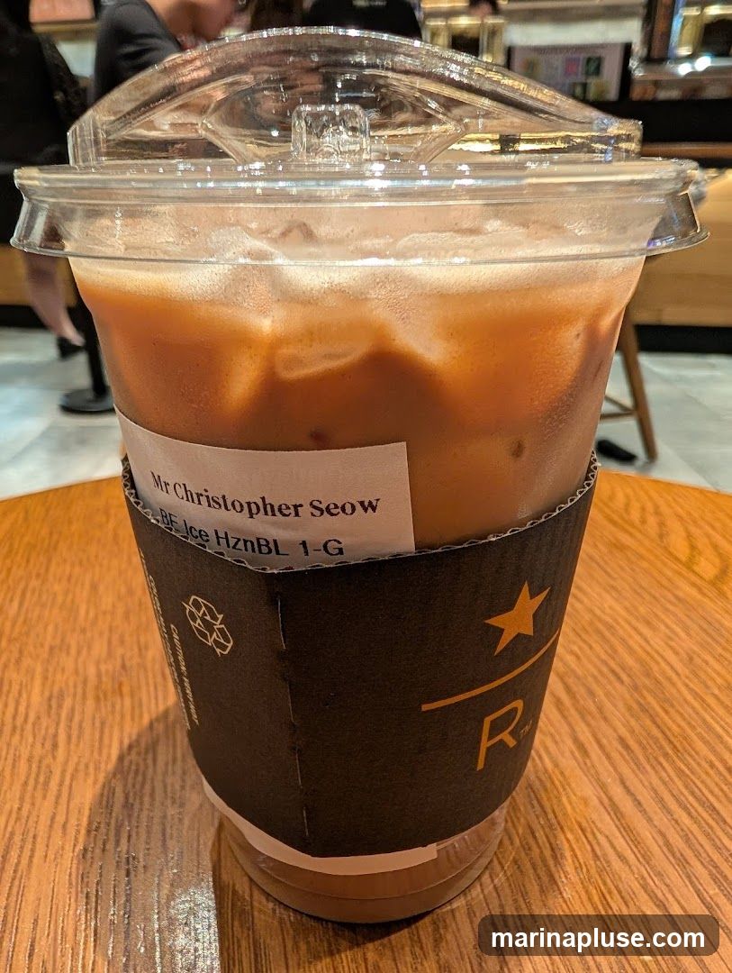 A close-up shot of an iced coffee in a clear plastic cup, placed on a wooden table. The cup has a cardboard sleeve with a logo and a white label that reads "Mr Christopher Seow" and "BE Ice HznBL 1-G". The drink appears to be a blend of coffee and milk, with ice cubes visible. In the background, blurred figures and shelves suggest an indoor setting, possibly a cafe.
