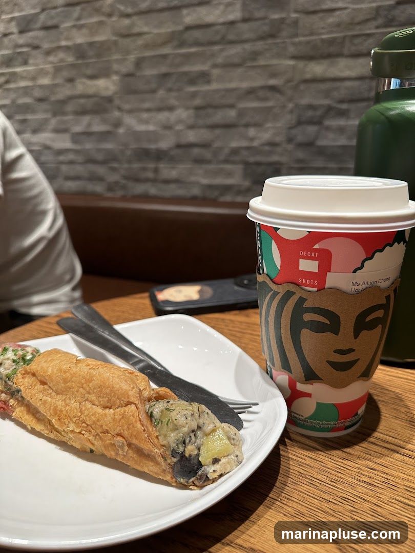 A close-up shot of a Starbucks coffee cup and a sandwich on a white plate, placed on a wooden table. The coffee cup has a festive holiday design and the iconic Starbucks siren logo. The sandwich appears to be a long roll filled with a mixture of ingredients. A green water bottle is visible in the background, along with a person's shoulder and arm. The wall behind the table is decorated with a textured stone pattern.