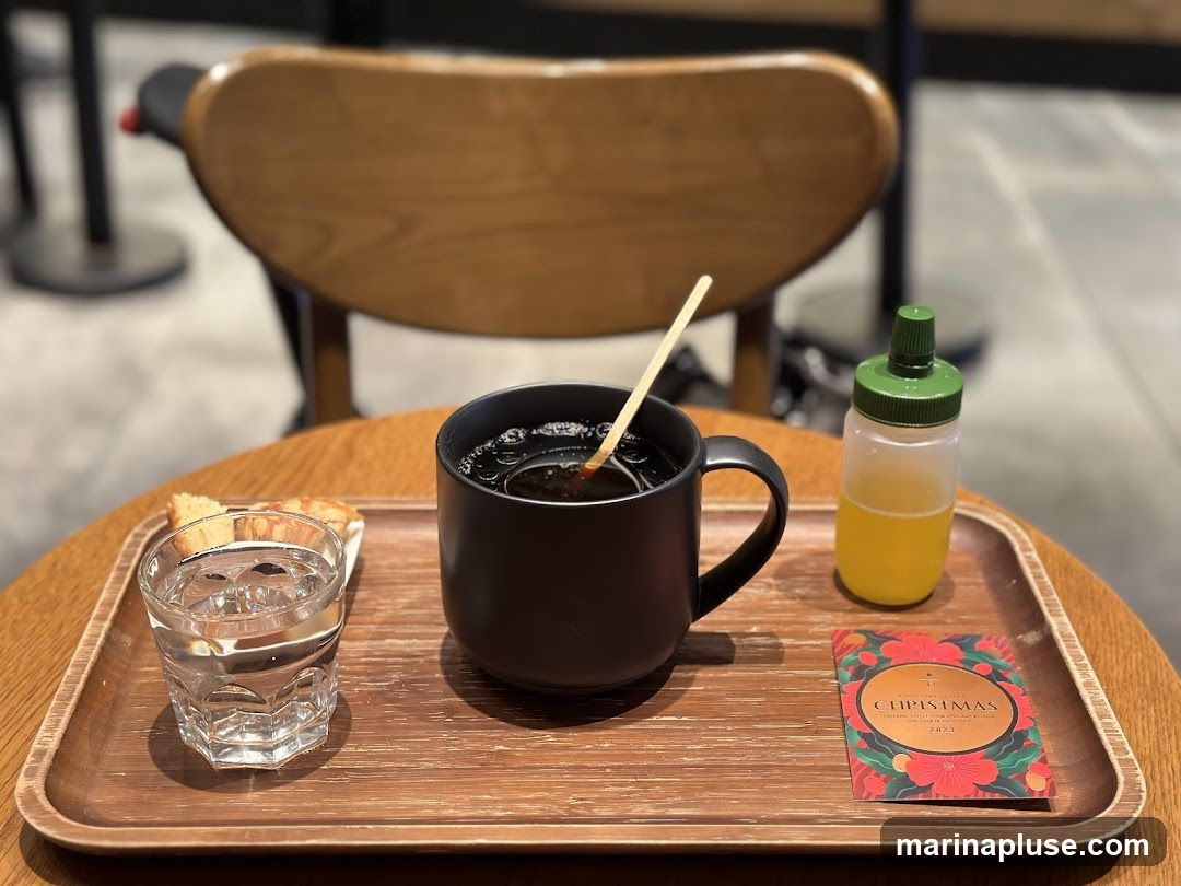 A black mug of coffee with a wooden stirrer, a glass of water with a piece of pastry, and a small bottle of yellow liquid are arranged on a wooden tray. A festive card with "CHRISTMAS 2023" is also on the tray. The setting appears to be a cafe with wooden chairs and tables.