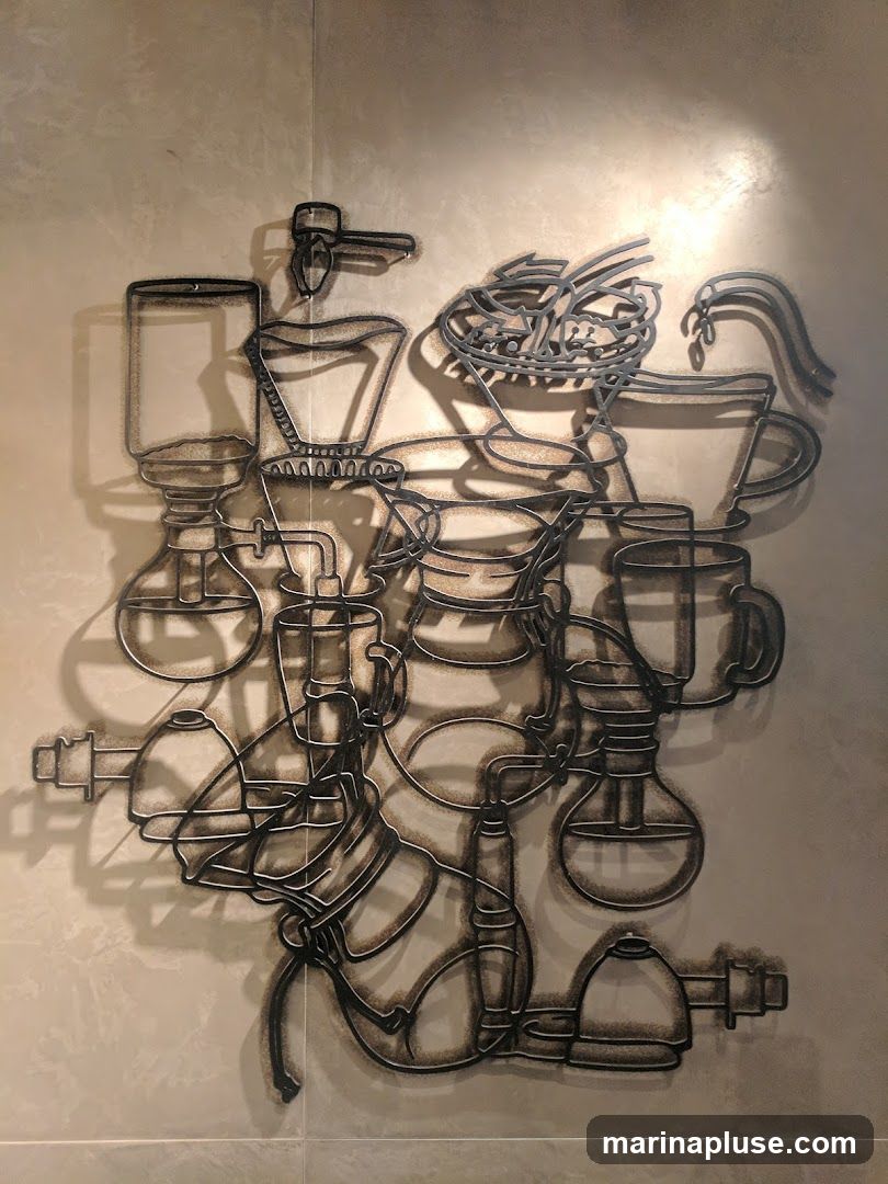 A metal wire sculpture depicting various coffee brewing apparatus, including pour-over devices, grinders, and cups, is mounted on a tiled wall. The artwork casts shadows on the wall, creating a dynamic visual effect.