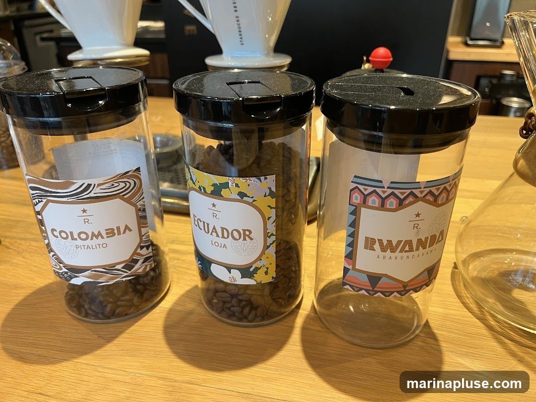 Three clear jars with black lids sit on a wooden surface. Each jar has a label indicating a coffee origin. The leftmost jar is labeled "COLOMBIA PITALITO" and contains coffee beans. The middle jar is labeled "ECUADOR LOJA" and also contains coffee beans. The rightmost jar is labeled "RWANDA ABAKUNDAKAWA" and is empty. In the background, white ceramic pour-over coffee drippers are visible.