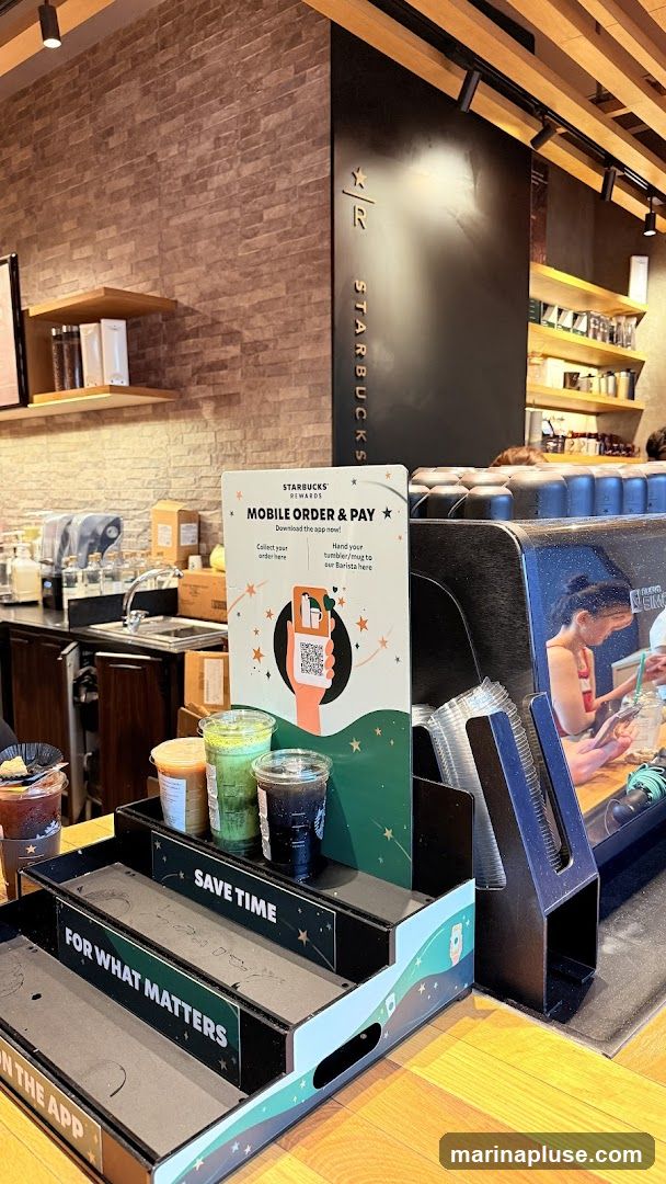 A display promoting Starbucks' mobile order and pay service is shown. It features instructions to download the app, collect orders, and hand in tumblers or mugs to the barista. Several drinks are displayed on tiered stands, including what appears to be iced coffee and matcha drinks. The Starbucks logo is visible on the back wall.