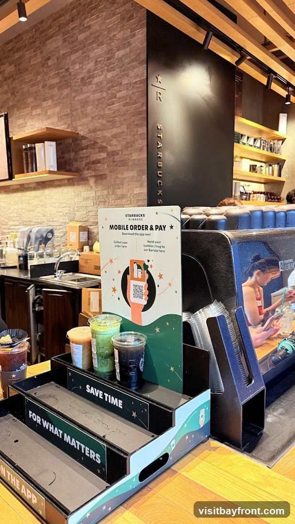 A display promoting Starbucks' mobile order and pay service is shown. It features instructions to download the app, collect orders, and hand in tumblers or mugs to the barista. Several drinks are displayed on tiered stands, including what appears to be iced coffee and matcha drinks. The Starbucks logo is visible on the back wall.