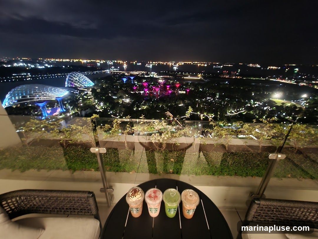 Four drinks are placed on a round table on a balcony. The drinks appear to be iced beverages with whipped cream and toppings. In the background, a city skyline is visible at night, with illuminated buildings and lights stretching into the distance. The view includes a large park with glowing structures.