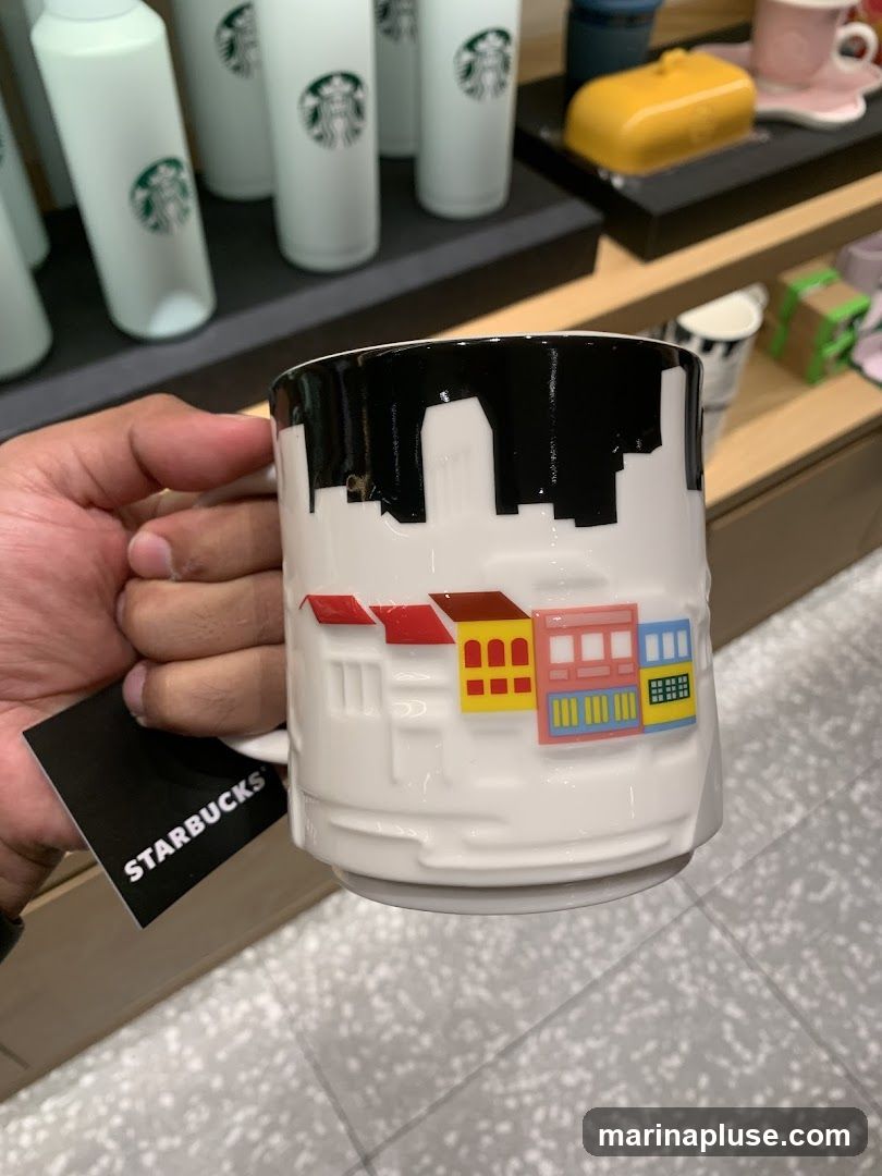 A hand holds a white Starbucks mug decorated with a black cityscape silhouette at the top and colorful, embossed buildings below. In the background, several white Starbucks tumblers with green logos are displayed on shelves, along with other merchandise. A black tag with the white Starbucks logo is visible on the mug.