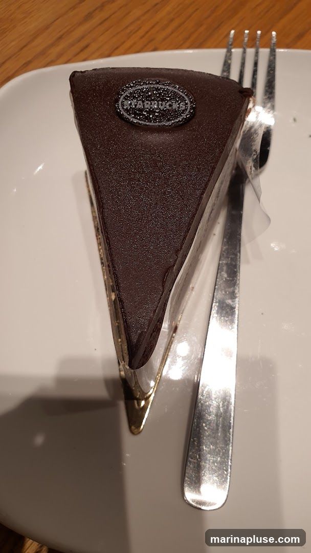 A slice of chocolate cake with a Starbucks logo on top, served on a white plate with a fork. The cake has a dark chocolate glaze and appears to be layered. The background is a wooden table.