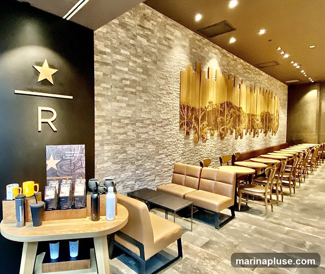 The interior of a Starbucks cafe with a prominent Starbucks logo on the left wall. The cafe features a long row of tables and chairs, as well as comfortable seating areas with sofas and armchairs. A decorative mural depicting a cityscape or landscape adorns the stone-tiled wall. On a wooden display table, there are coffee bags, mugs, and reusable water bottles.