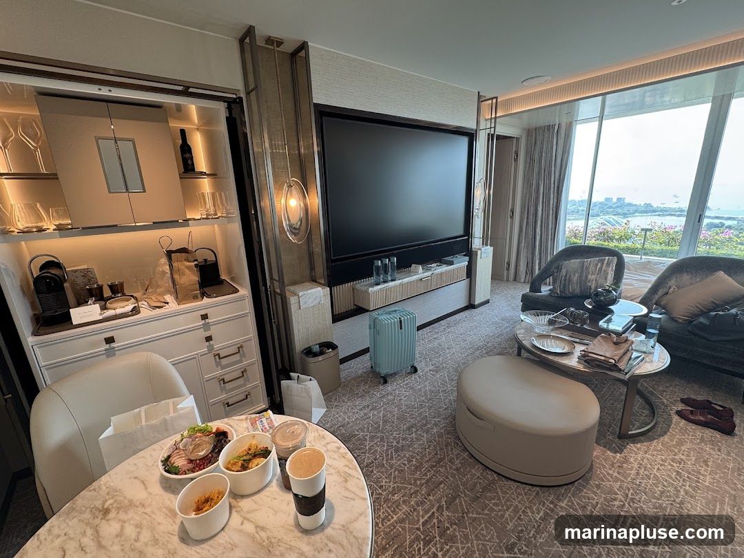 A hotel room with a large television, a minibar stocked with drinks and coffee machine, and a dining table set with takeaway food. There are two armchairs and an ottoman in the seating area, and a large window offers a view of the city and water.