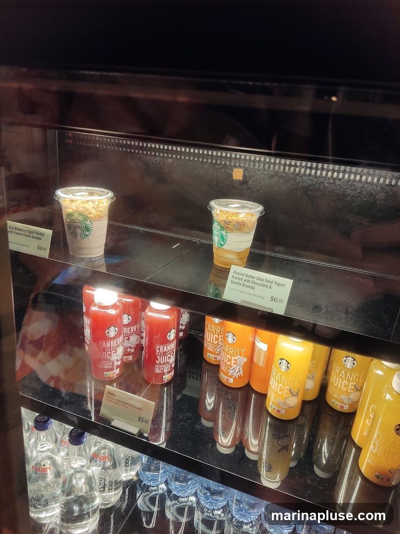 A refrigerated display case filled with various beverages and parfaits. On the top shelf, there are two parfaits: an Acai Blueberry Yogurt Parfait with French Vanilla Granola priced at $6.00, and a Peanut Butter Chia Seed Yogurt Parfait with Chocolate & Vanilla Granola priced at $6.50. The middle shelf contains bottles of Cranberry Red Grape Juice and Carrot Juice, along with bottles of Orange Juice. A sign indicates "Juices" for $5.50. The bottom shelf holds bottles of Evian sparkling water and other bottled water.