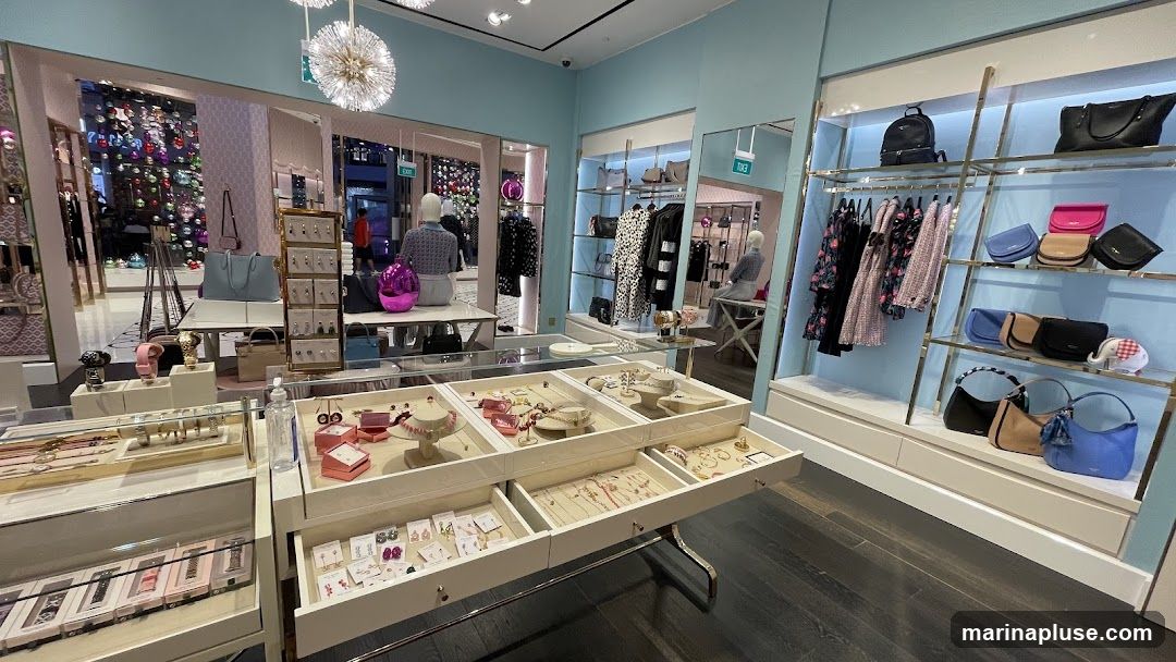 The interior of a brightly lit store displays a variety of merchandise, including handbags, clothing, watches, and jewelry. The store features light blue walls and white display units. In the foreground, a long display case showcases necklaces, earrings, and bracelets. To the right, shelves are stocked with colorful handbags and a decorative elephant figurine. Racks of clothing, including dresses and jackets, are visible in the background. Mirrors and mannequins are also present.