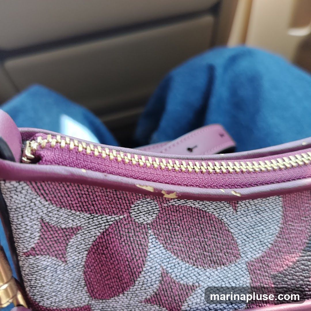 A close-up view of a handbag with a pink and white pattern. The zipper is visible, and the strap is a light pink color. The background is blurred, showing what appears to be blue fabric and a tan surface.