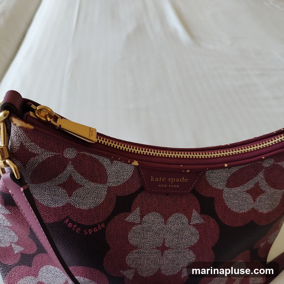 A close-up of a Kate Spade handbag with a floral pattern. The bag is burgundy with a repeating pattern of stylized flowers in shades of pink, white, and gray. The bag has gold-toned hardware, including a zipper and a small tag with the Kate Spade New York logo. The strap is visible on the left side of the image.