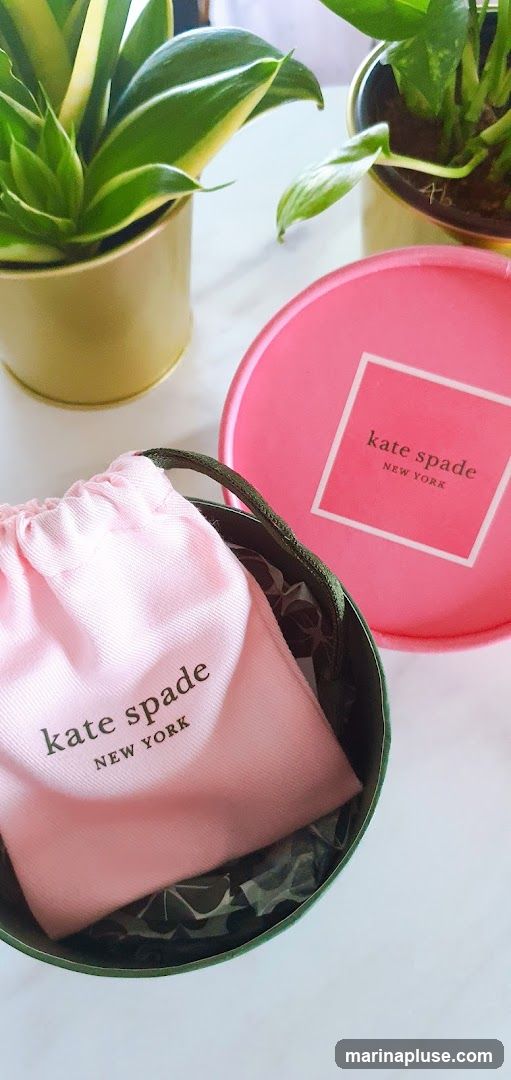A pink drawstring pouch and a pink circular box, both branded with "kate spade NEW YORK", are placed on a white surface next to two potted plants. The pouch is inside a dark green cylindrical container.