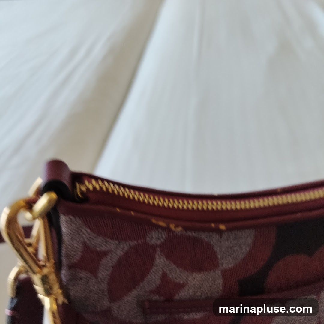 A close-up shot of a handbag with a repeating pattern of maroon and grey circles. The top edge of the bag features a gold zipper and maroon trim. A gold clasp and strap are visible on the left side. The background is a blurred white fabric.