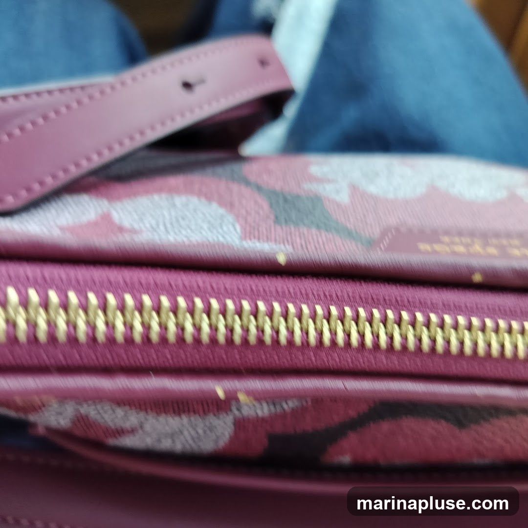 A close-up shot focuses on the textured details of a burgundy bag with a floral pattern in shades of pink, gray, and black. A prominent gold zipper runs across the middle of the frame, with its teeth clearly visible. A strap, also in burgundy, is visible at the top left, with a button closure. The background consists of blurred denim fabric. The brand name "BYRON" is faintly visible in gold lettering on the bag's surface.