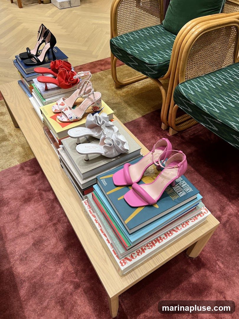 A collection of women's high-heeled shoes are artfully arranged on a stack of books placed on a light wood coffee table. The shoes include black open-toe heels, red ruffled sandals, white ruffled slides, floral-patterned sandals, and bright pink ankle-strap heels. The scene is set in a well-lit room with rattan chairs featuring green patterned cushions visible in the background, and a plush rug on the floor.