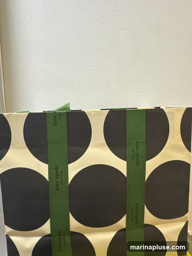 A shopping bag with a black and cream polka dot pattern. Green ribbon handles are visible, with the "kate spade NEW YORK" logo printed vertically on them.