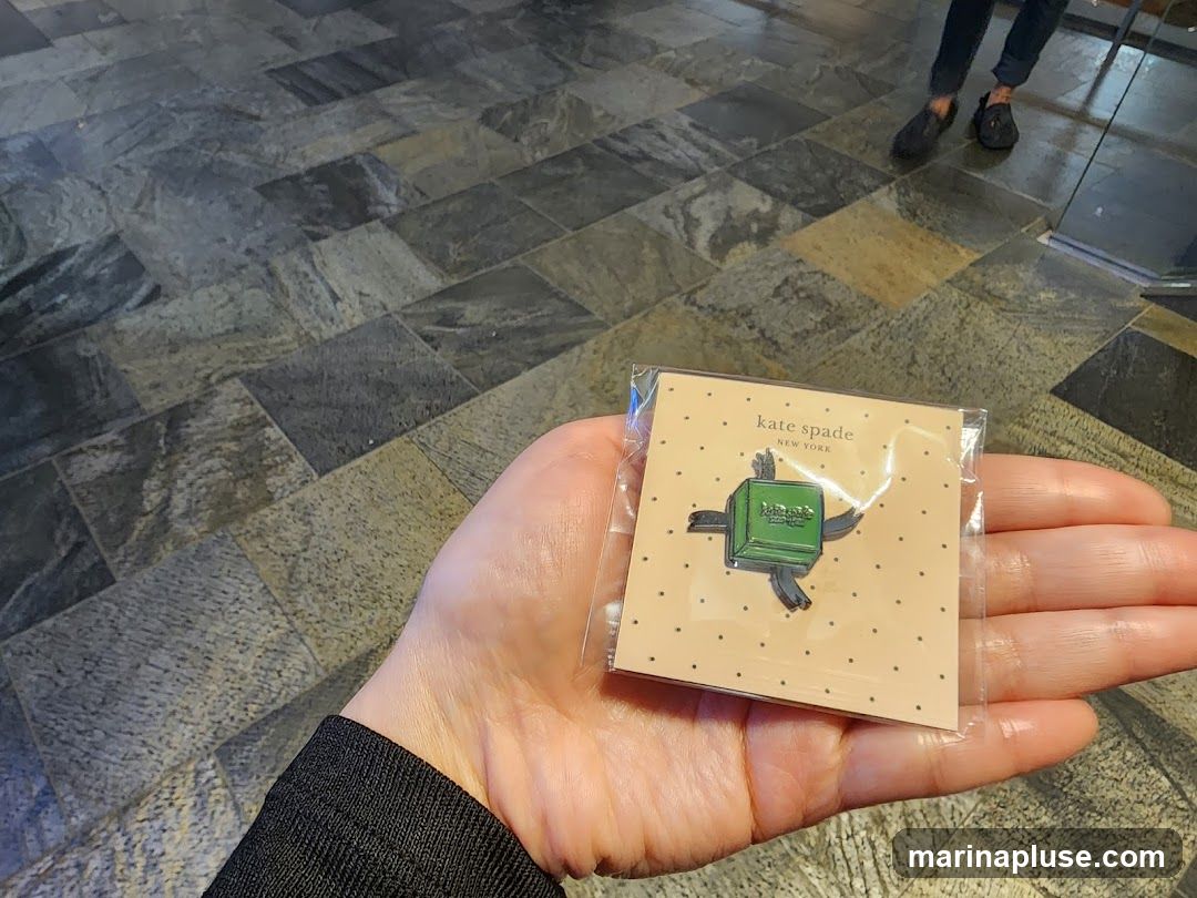 A hand holds a packaged Kate Spade New York pin featuring a green turtle. The pin is on a light brown card with small black dots. The background is a tiled floor.