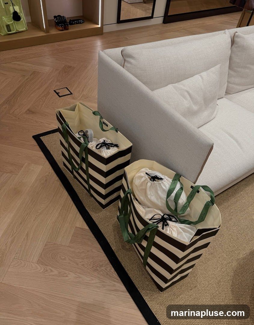 Two striped shopping bags with green handles are placed on a rug next to a light-colored sofa. The bags contain white drawstring pouches. The floor is made of light wood in a herringbone pattern. In the background, a shelf displays shoes and a green bag.