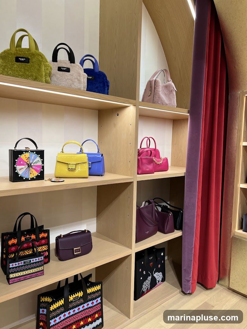 A retail display featuring various handbags on wooden shelves. The bags include fuzzy textured styles in green, cream, and blue, as well as patent leather bags in yellow and blue. There are also structured bags in black, pink, and burgundy, and tote bags with colorful embroidered patterns and elephant designs. A pink fuzzy handbag is visible on the top right shelf. Red and purple velvet curtains hang to the right of the display.