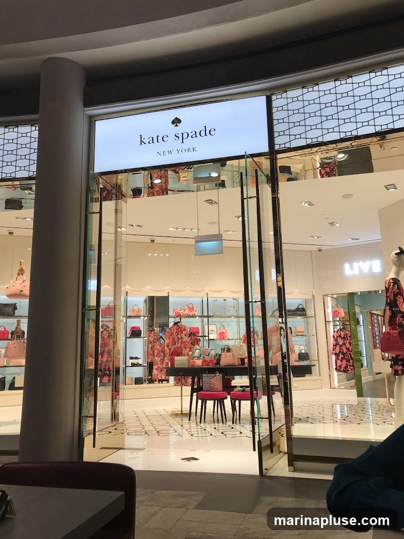The exterior of a Kate Spade New York store. The store has a large illuminated sign with the brand name and "New York" below it. The storefront features a glass facade with a display of handbags, clothing, and accessories. Inside, mannequins are dressed in floral print outfits, and shelves are stocked with various bags. A small seating area with a table and pink stools is visible near the entrance.