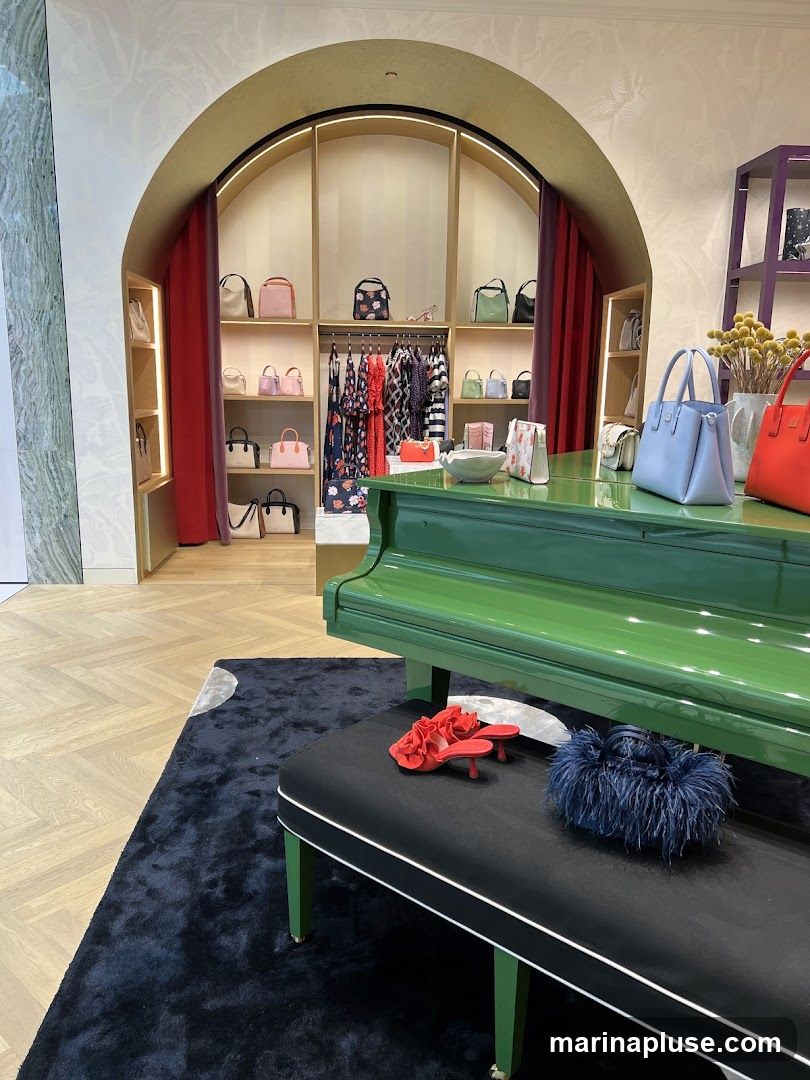 A retail store interior featuring a display of handbags and clothing. A green grand piano serves as a display surface for several handbags, including a light blue one and a red one. A black bench in front of the piano holds a pair of red ruffled heels and a dark blue feathered handbag. Behind the piano, shelves are stocked with various handbags in different colors and styles. A rack of clothing, including dresses and tops, is also visible. The store has a modern and elegant aesthetic with light-colored walls and wooden flooring.