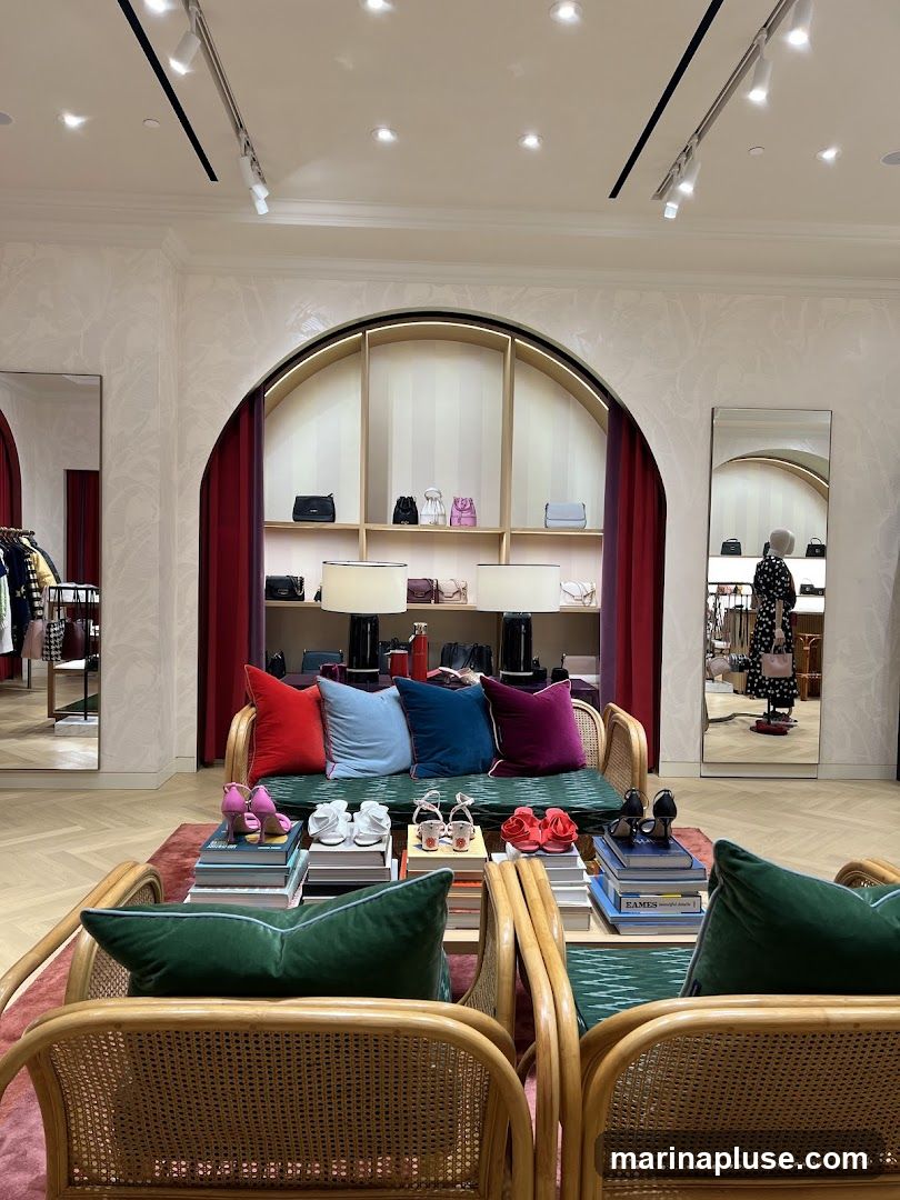 The image shows a well-lit, luxurious store interior. In the foreground, there are two rattan chairs with green velvet cushions. Between them is a coffee table displaying stacks of books topped with various shoes and decorative items. Behind the coffee table is a green sofa with colorful velvet cushions (red, light blue, dark blue, and purple). A large arched display unit with shelves filled with handbags is visible in the background, framed by red curtains. Full-length mirrors are placed on either side of the display unit, reflecting the store's ambiance and merchandise. Racks of clothing and more handbags are visible in the distance.