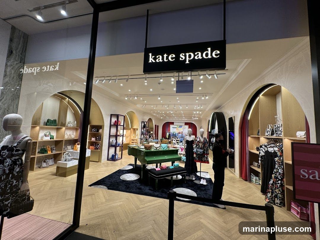 A view into a Kate Spade store through a large window. The store features a green grand piano in the center, surrounded by displays of handbags and clothing. Mannequins are dressed in patterned outfits. The store's name, "kate spade", is prominently displayed on a black sign above the entrance. The interior has arched displays and warm lighting. A sign with the word "sale" is visible on the right.