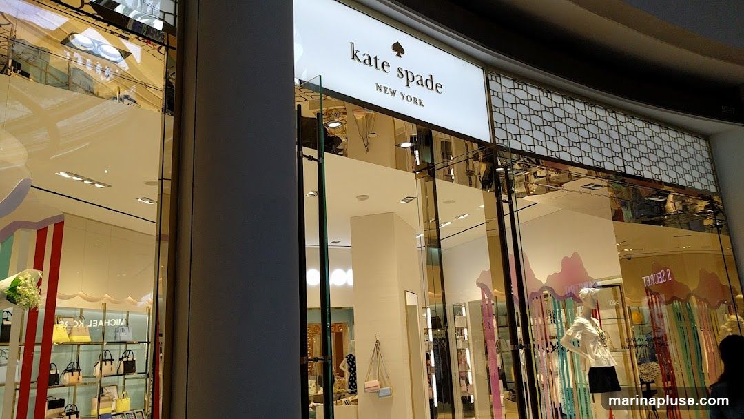 The exterior of a Kate Spade New York store. The store has a large illuminated sign with the brand name and logo. The storefront features large glass windows displaying merchandise, including handbags and clothing on mannequins. The interior is visible through the windows, showing a well-lit retail space.