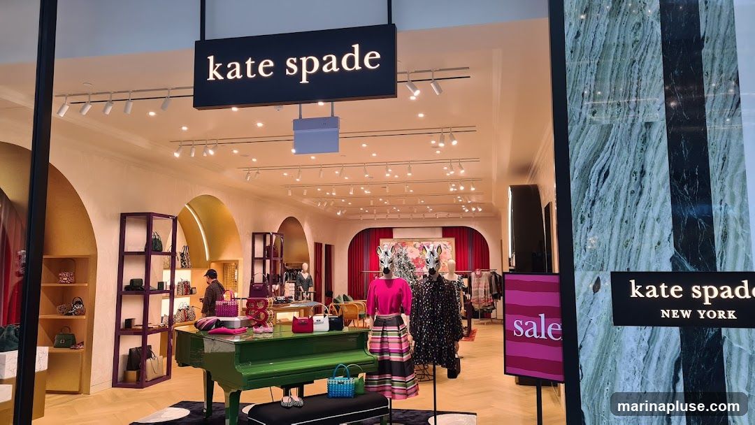 The exterior of a Kate Spade store is visible, with the brand's name displayed prominently on a black sign above the entrance. Inside, the store is brightly lit and showcases handbags, clothing on mannequins, and decorative elements like a green piano. A "sale" sign is visible to the right.