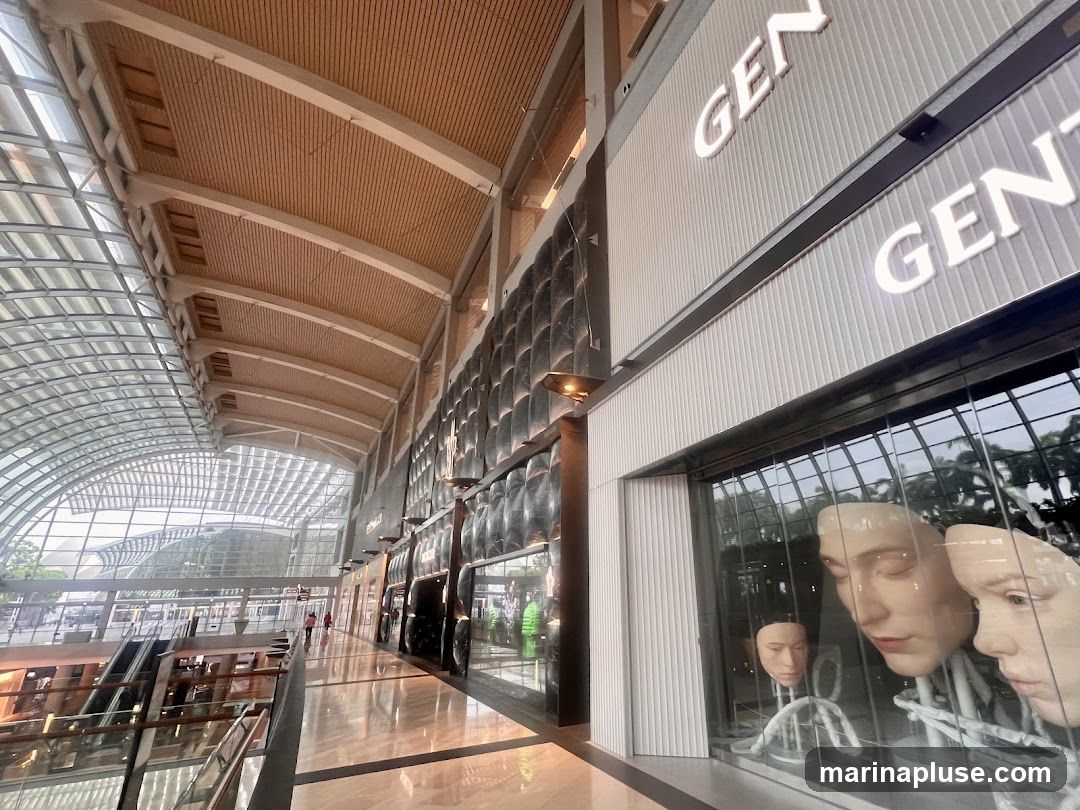 A modern store facade with the word "GEN" displayed in illuminated letters. The storefront window displays several realistic mannequins with distinct facial features. The interior of the store is visible through the window, and the overall aesthetic is sleek and contemporary. The background shows a large, airy atrium with a distinctive arched ceiling and glass walls, suggesting a shopping mall or airport terminal.