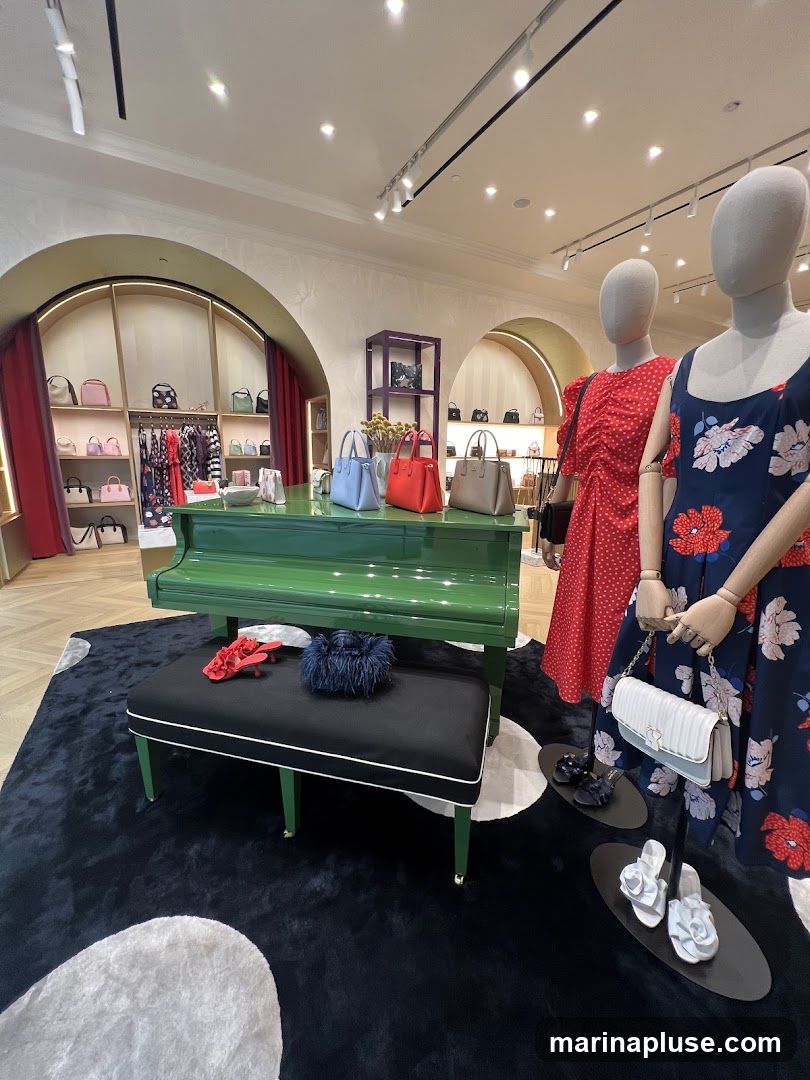 A retail store interior features a display with mannequins dressed in floral print dresses and a red polka dot dress. A bright green piano serves as a central display piece, adorned with several handbags in various colors. A black bench with white trim sits in front of the piano, holding a pair of red heels and a fluffy blue clutch. The floor is covered with a dark rug featuring large white polka dots. Shelving units in the background showcase a collection of handbags and clothing items.