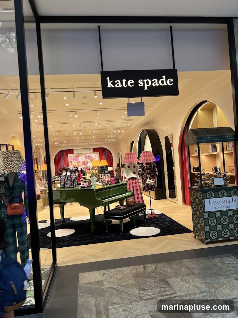 The exterior of a Kate Spade store. A black sign with white lettering that reads "kate spade" is visible above the entrance. Inside, the store is decorated with a green grand piano, mannequins displaying clothing, and various accessories.