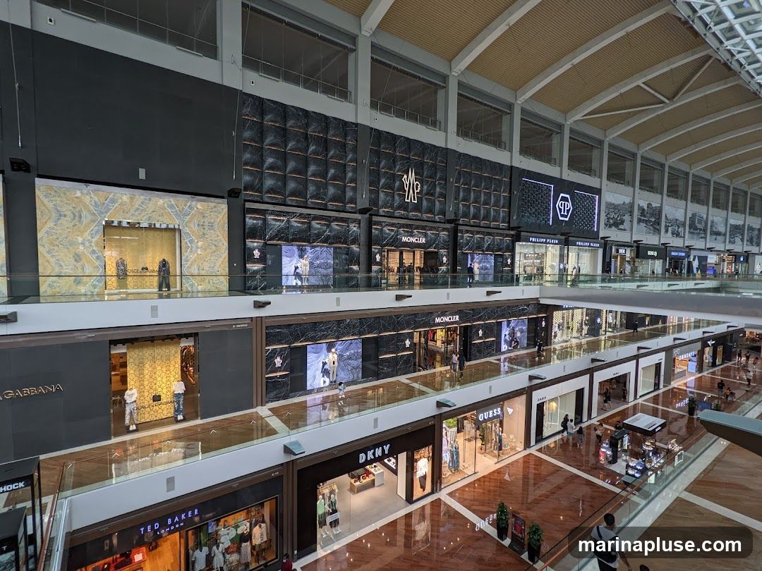 An interior view of a multi-level luxury shopping mall with various high-end stores. Visible brands include Dolce & Gabbana, Moncler, Philipp Plein, DKNY, Guess, and Ted Baker. The architecture features a modern design with a high ceiling, glass railings, and polished floors. Mannequins are displayed in store windows, showcasing fashion items. Shoppers are seen browsing on different levels.