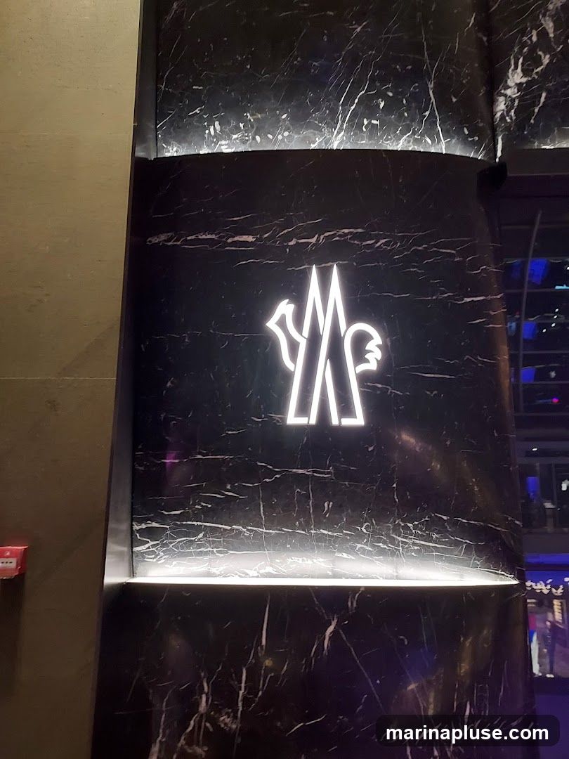 A close-up shot of the Moncler logo illuminated in white neon against a black marble-patterned wall. The logo is a stylized representation of a rooster. The wall has a polished finish with white veining.