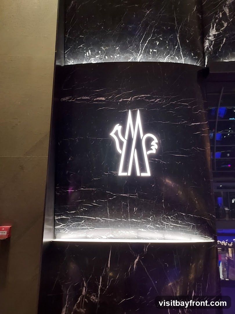 A close-up shot of the Moncler logo illuminated in white neon against a black marble-patterned wall. The logo is a stylized representation of a rooster. The wall has a polished finish with white veining.