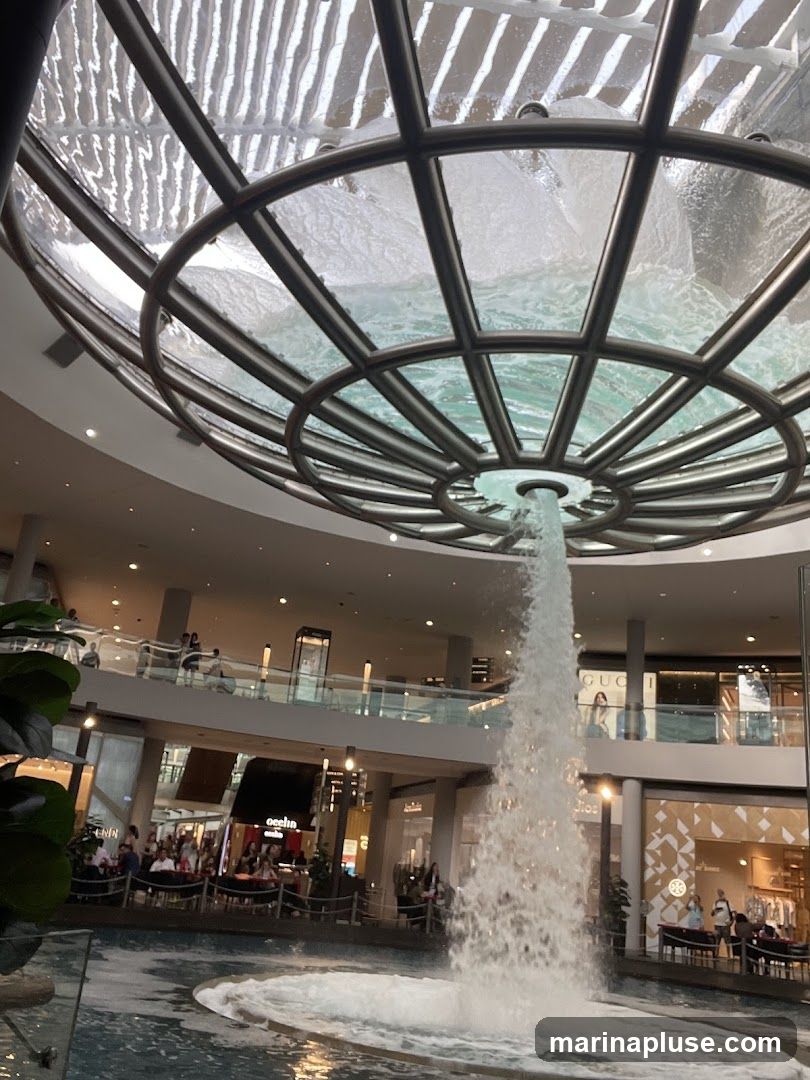 A large indoor fountain cascades water from a circular structure in the ceiling of a modern shopping mall. The mall features multiple levels with shops and seating areas visible in the background.