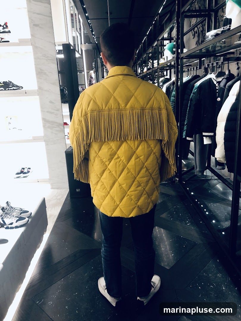 A person stands in a retail store, facing away from the camera. They are wearing a yellow quilted jacket with fringe detailing on the back and sleeves, dark wash jeans, and white sneakers. The store interior is visible, with shelves displaying shoes and racks of clothing in the background.