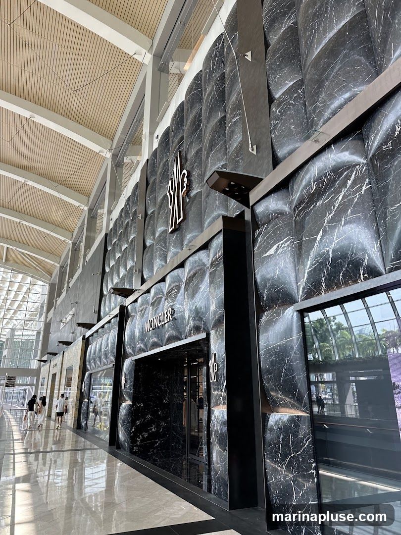 The exterior of a Moncler store with a black marble facade. The Moncler logo is prominently displayed above the entrance. The store is located in a modern indoor shopping area with a high ceiling and natural light.