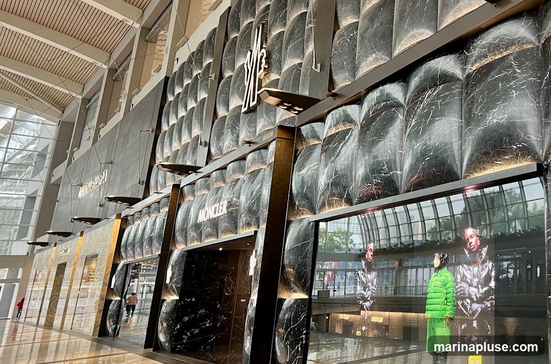 The exterior of a Moncler store with a distinctive marble-patterned facade. The Moncler logo is prominently displayed above the entrance. Inside, mannequins are visible, showcasing puffer jackets.