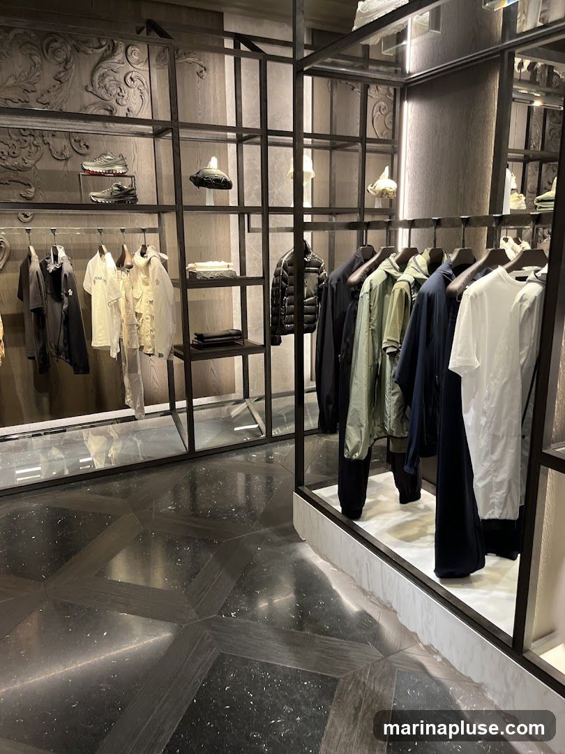 A well-lit clothing store interior featuring a display of men's apparel. Racks of jackets, shirts, and pants are neatly arranged on shelves and hanging rails. Several pairs of sneakers are displayed on upper shelves. The flooring is a dark, polished stone with a geometric pattern. The overall aesthetic is modern and minimalist.