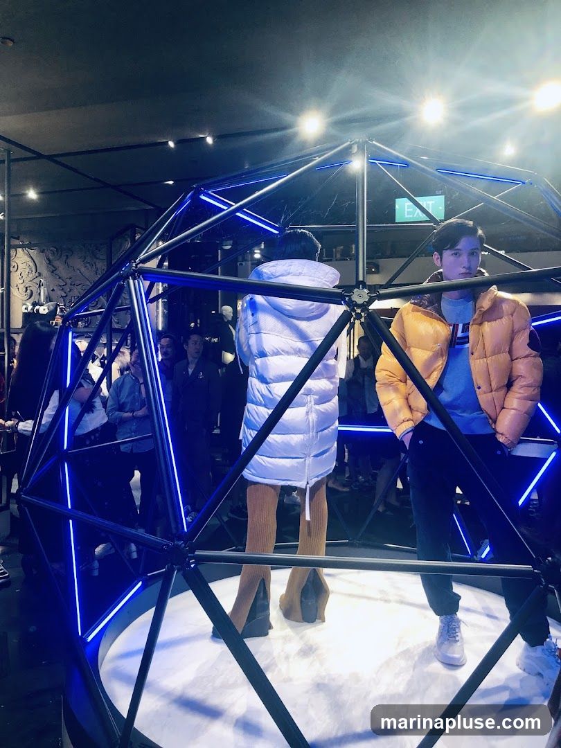 A fashion display featuring mannequins wearing puffer jackets inside a geodesic dome structure with blue LED lighting. One mannequin wears a white puffer jacket and brown ribbed leggings, while another wears an orange puffer jacket. Several people are visible in the background, observing the display. An "EXIT" sign is visible in the upper right portion of the image.