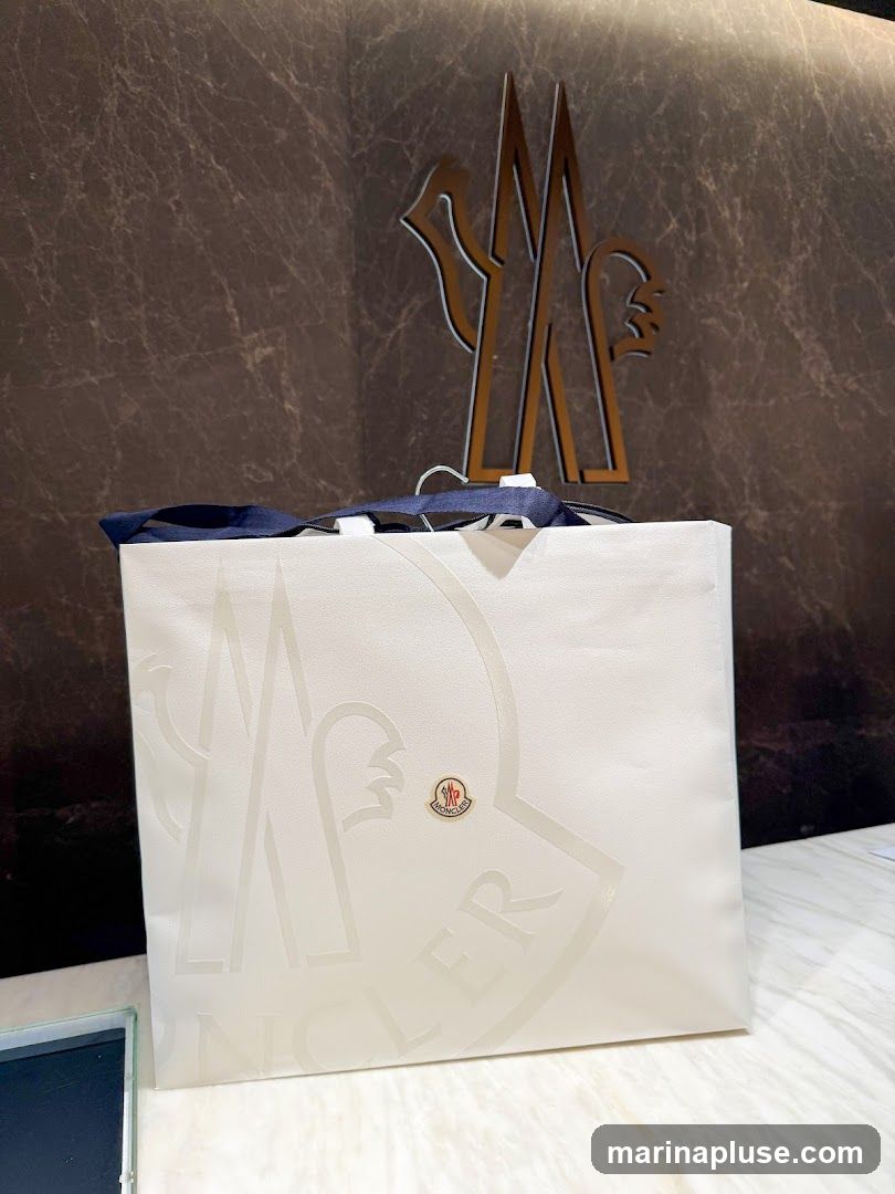 A white Moncler shopping bag with the brand's logo embossed on it. A smaller Moncler logo badge is also visible on the bag. In the background, a large Moncler logo is mounted on a dark, marble-patterned wall.