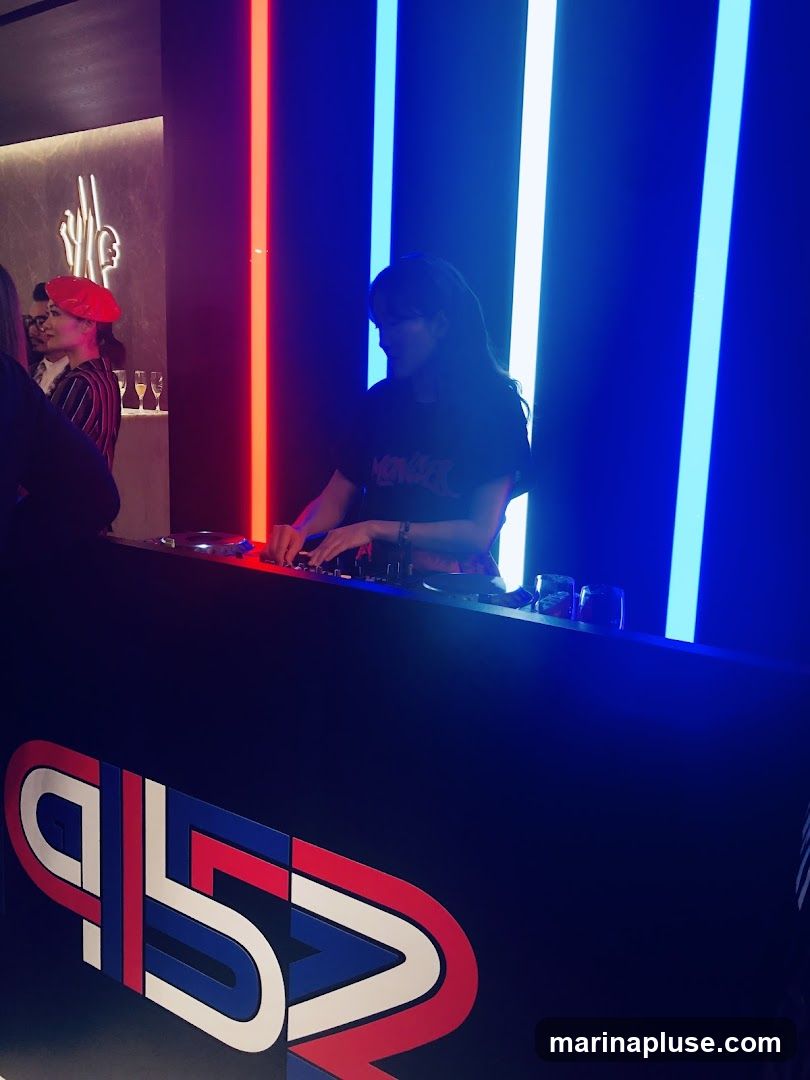 A DJ is seen playing music at a counter with a large, colorful logo that reads "915". The background features vertical neon lights in red, white, and blue. To the left, a person wearing a red beret and a striped shirt is visible, along with a bar area with champagne flutes. A Moncler logo is subtly displayed on the wall in the background.