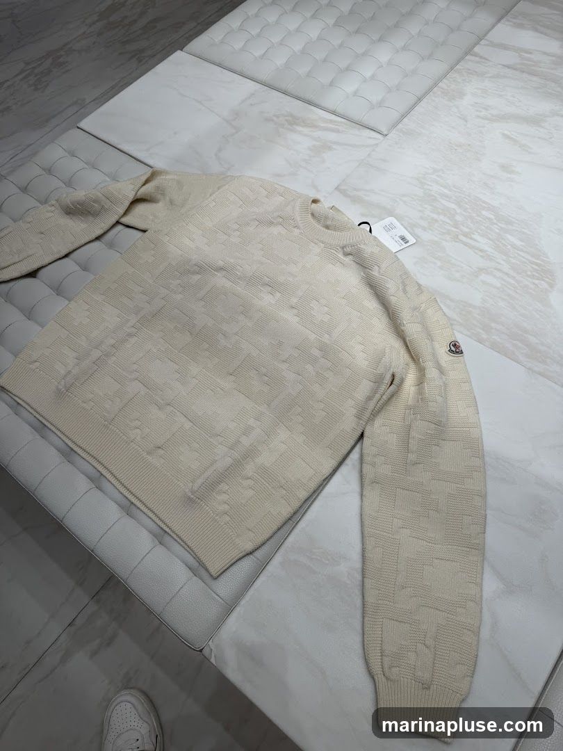 A cream-colored Moncler sweater with a textured geometric pattern is laid out on a white tufted bench. A Moncler logo patch is visible on the left sleeve. A price tag is attached to the sweater. A white sneaker is visible in the lower left corner.