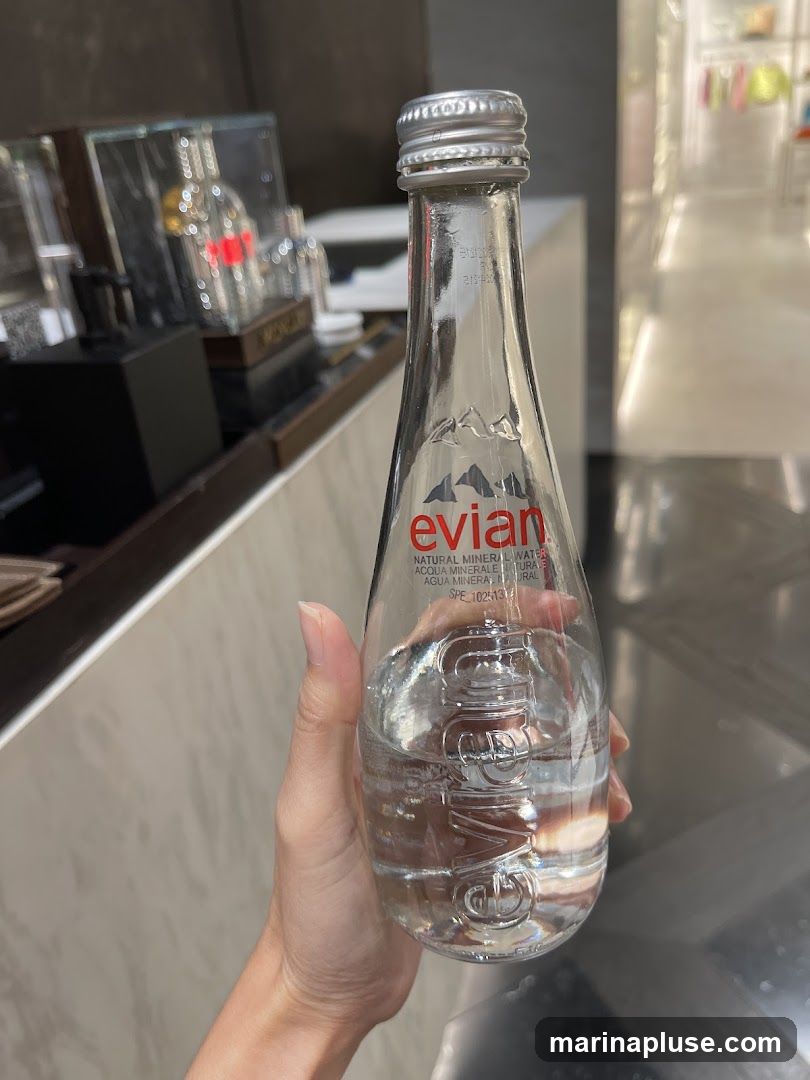 A hand holds a clear glass bottle of Evian natural mineral water. The bottle has a silver cap and the Evian logo with mountains is visible in red. The background shows a blurred interior of a store with displays and clothing racks.
