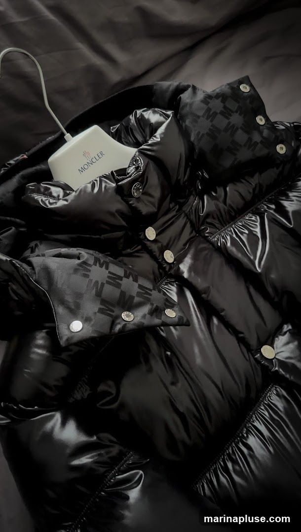 A close-up shot of a black, shiny Moncler puffer jacket. The jacket features a repeating pattern of the letter 'M' on some of its fabric panels. A white Moncler branded hanger is visible, holding the jacket.